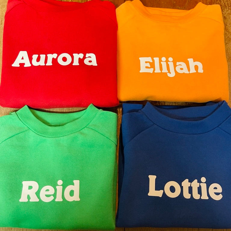 Four folded sweatshirts with names printed on them, arranged in a 2x2 grid on a wooden surface.