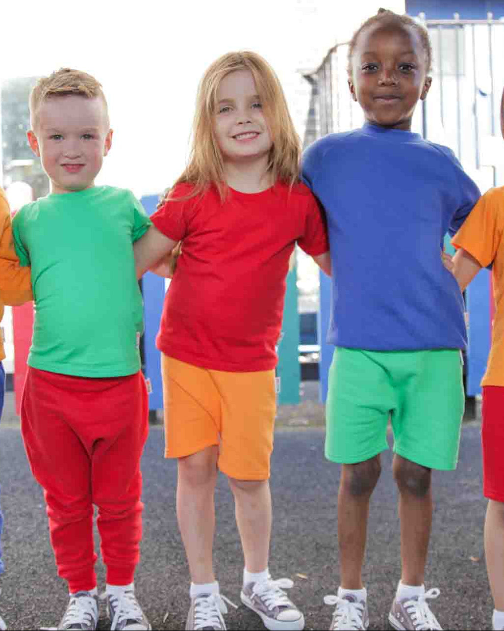 Kids in a playground wearing Hues Clothing standing in line with their arms around each other. 