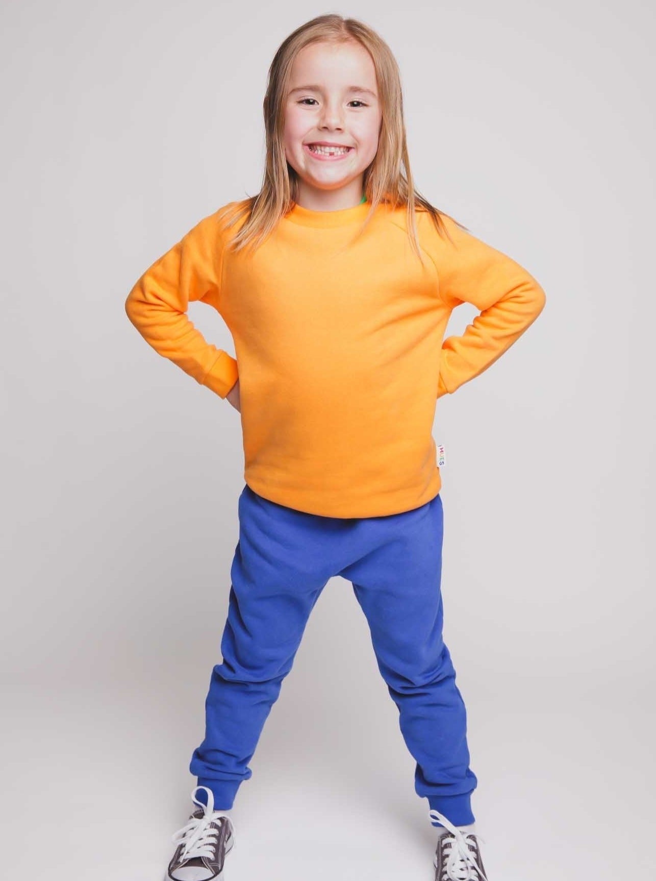 A blonde haired girl wearing an orange jumper and blue joggers - Hues Clothing